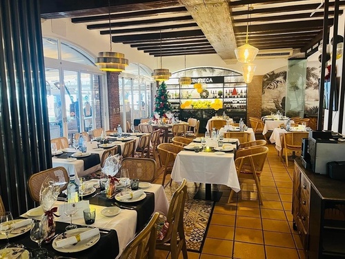 Se observa el interior de un restaurante con mesas elegantemente puestas para comer, adornado con motivos festivos y una vinoteca al fondo.