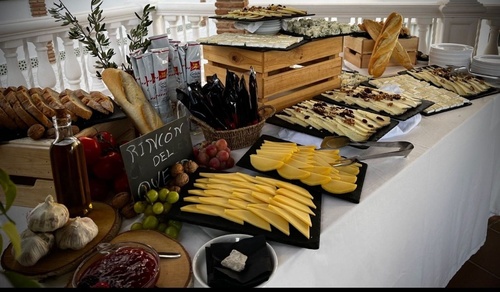 Una mesa presenta un abundante y variado bufé de quesos con pan, uvas, nueces y otros acompañamientos.