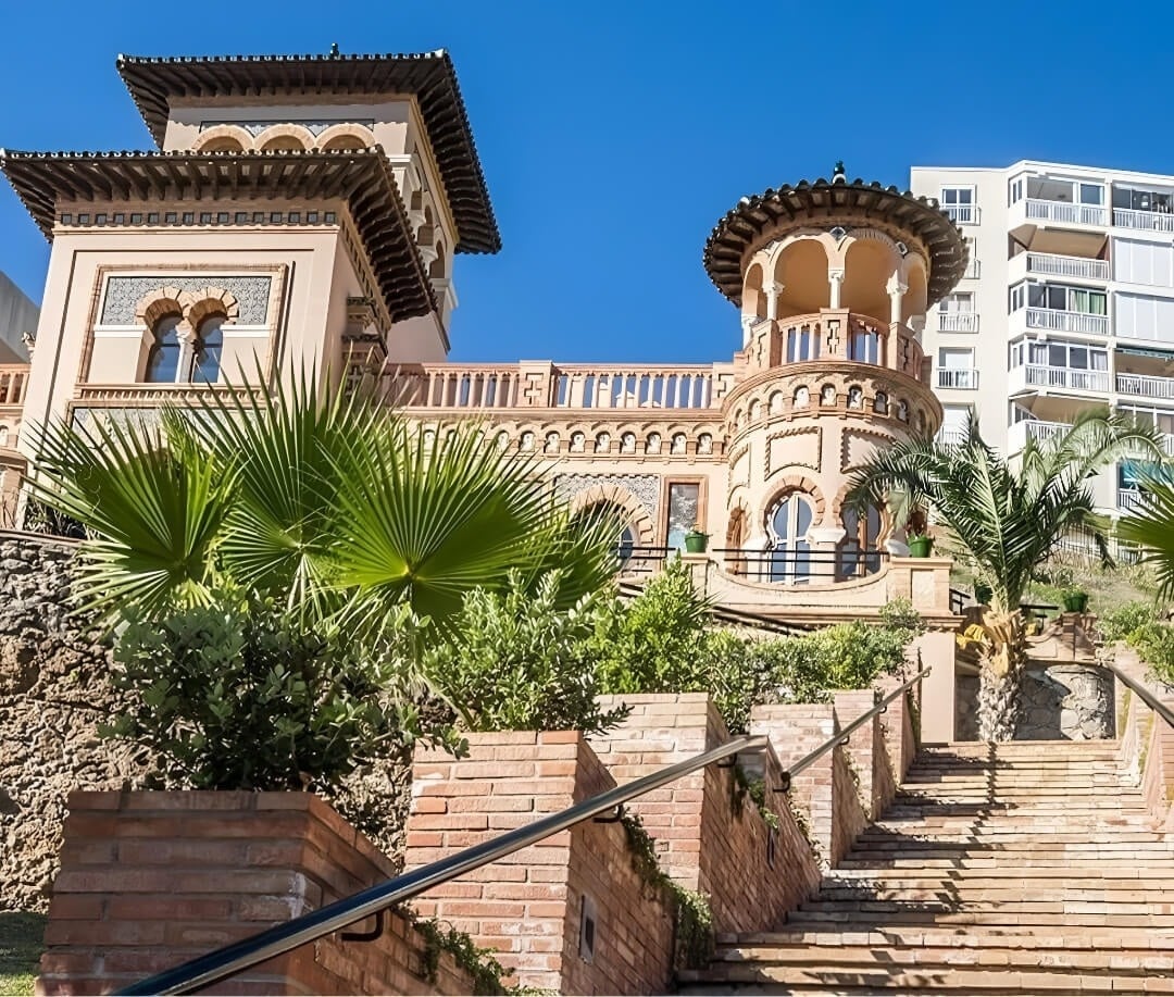 La imagen muestra un edificio de arquitectura ornamentada con torres y balcones, de estilo mediterráneo o morisco, rodeado de palmeras y vegetación, con una escalera de ladrillo en primer plano y un edificio moderno al fondo bajo un cielo azul.