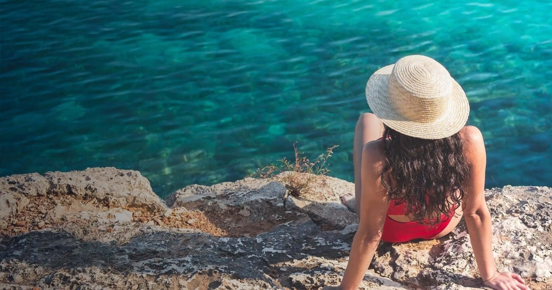 una mujer con traje de baño rojo y sombrero de paja se sienta en una roca junto al océano