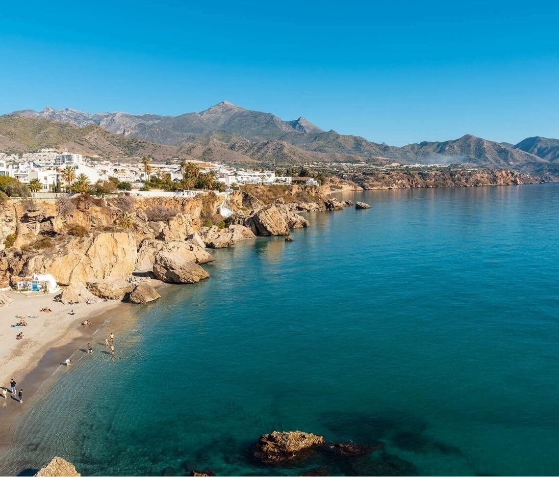 Una vista panorámica muestra un pueblo costero de casas blancas en acantilados rocosos, con una playa de arena y un mar azul turquesa, enmarcado por montañas bajo un cielo despejado.
