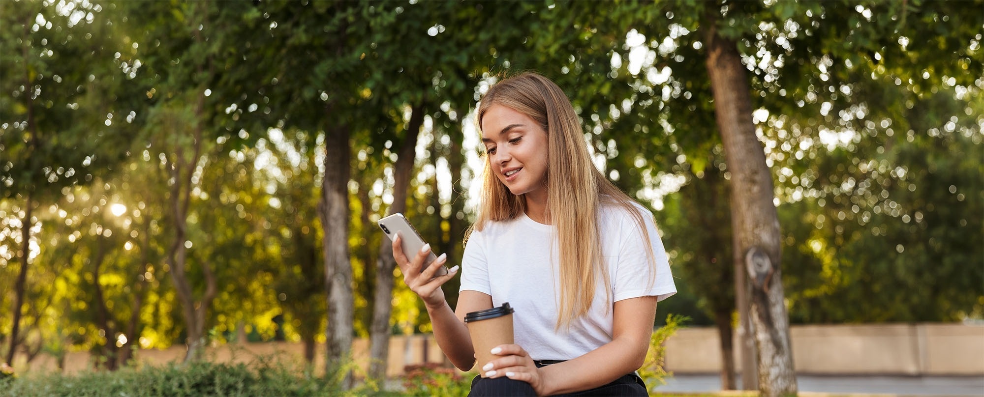 A young woman smiles while looking at her phone and holding a coffee cup outdoors amidst blurred trees and sunlight.