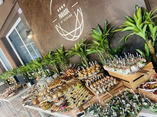 Una elaborada mesa de buffet exhibe una gran variedad de platillos y bocadillos elegantemente presentados para un evento, en un ambiente decorado con plantas y un logo de marca.
