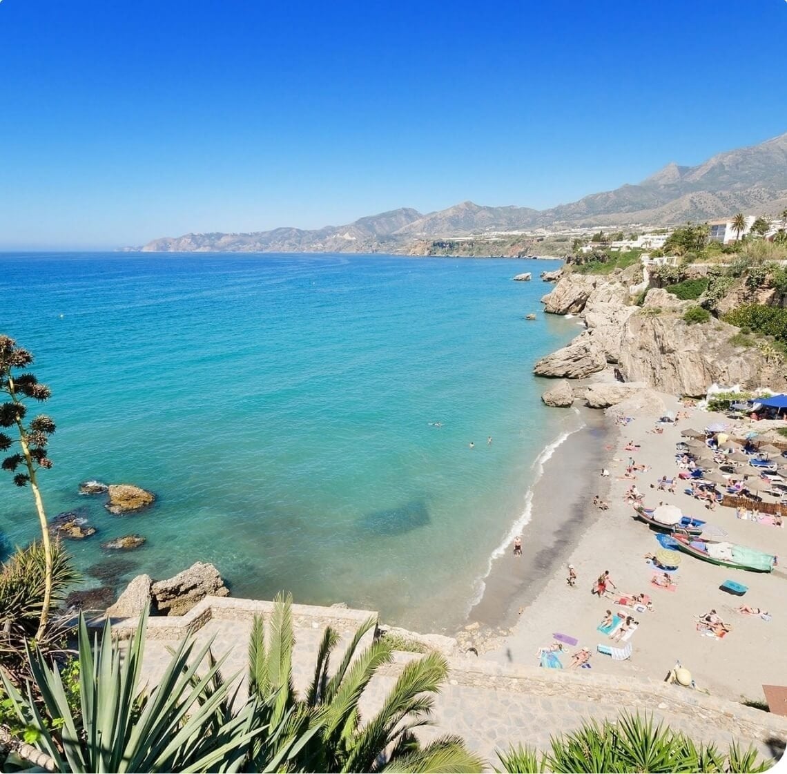 Vista elevada de una playa arenosa con bañistas, aguas turquesas, acantilados rocosos y montañas costeras bajo un cielo azul claro.