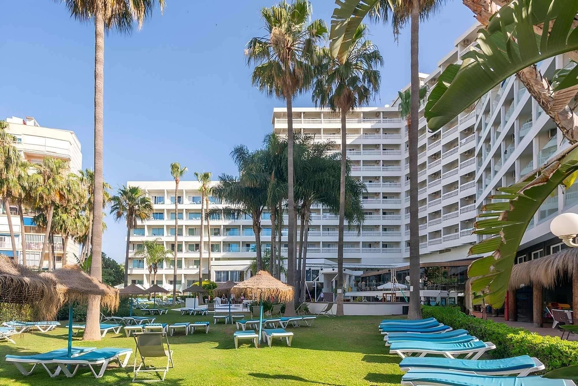 La imagen muestra un complejo hotelero con grandes edificios blancos, rodeado de palmeras y un césped verde con numerosas tumbonas y sombrillas de paja.