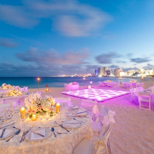 Beach wedding reception setup DEMO 3 at dusk, with illuminated dance floor and city skyline.