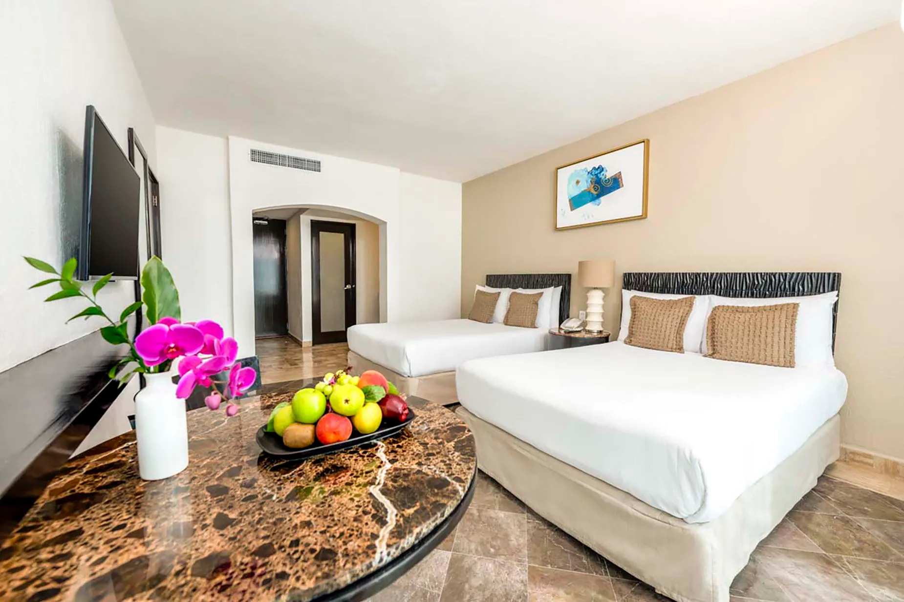 Two beds in a hotel room, with a fruit bowl and flowers on a marble table. DEMO 3