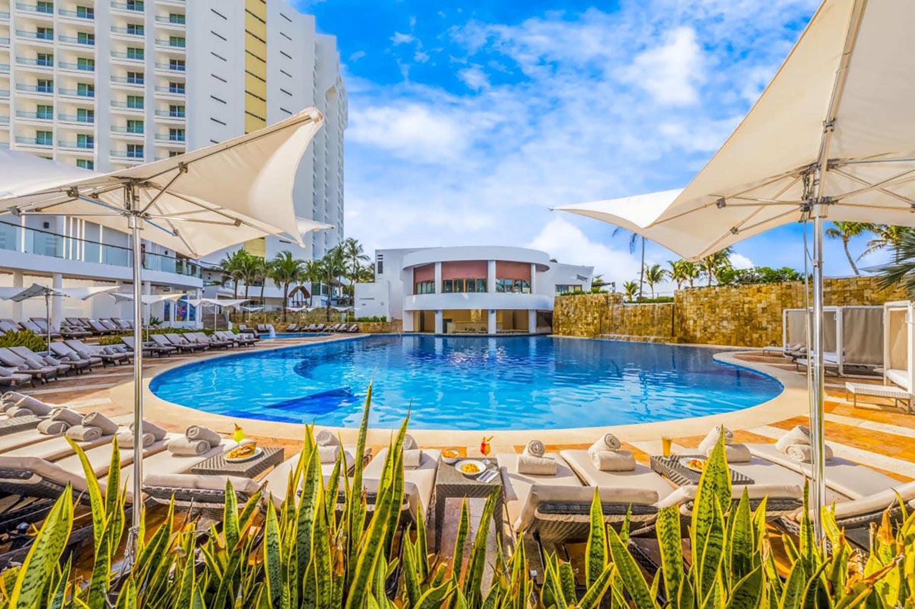 Crystal clear pool in front of hotel with lush greenery and sunbeds (Plantilla Baseline V6).