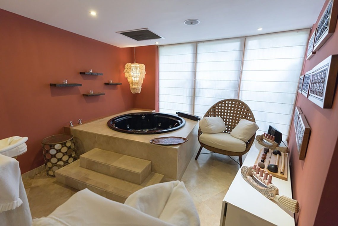 A warm-toned spa room features a built-in hot tub, a woven lounge chair, decorative shelving with candles, and large windows with privacy blinds.