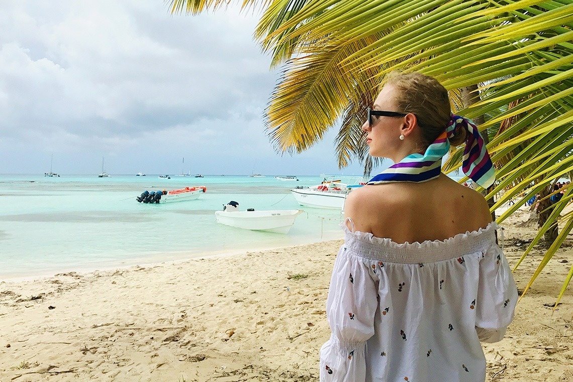 A woman wearing sunglasses and a colorful scarf looks out from a sandy beach at boats in clear turquoise water, beneath palm fronds and a cloudy sky.