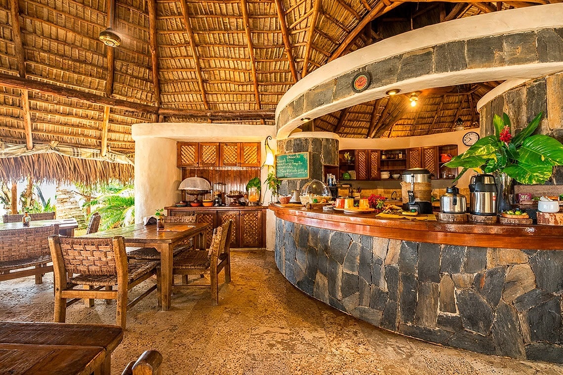 This inviting tropical dining area features a high thatched roof, rustic wooden furniture, and a stone-clad buffet counter laden with food and beverages.