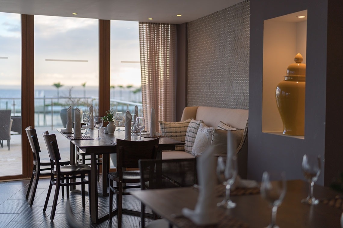 An elegant dining area overlooks the ocean through large windows, featuring wooden tables set with glassware and napkins, a comfortable banquette, and a decorative yellow jar in a wall alcove.