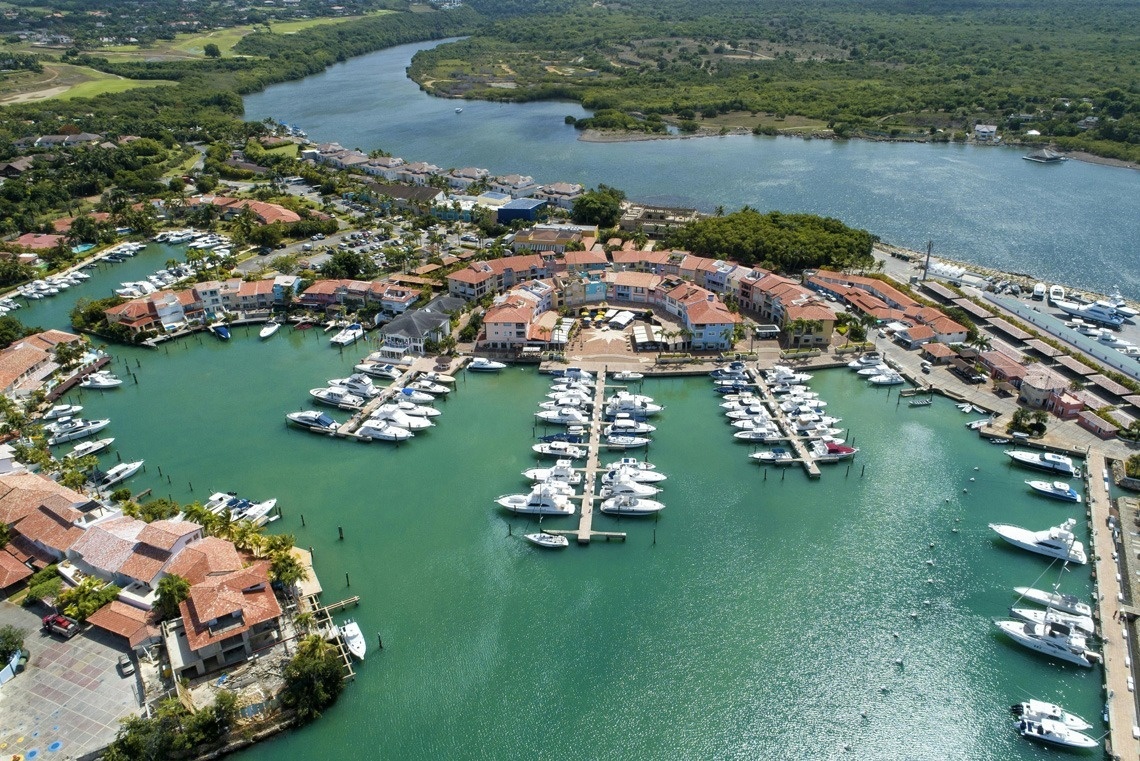 An aerial view captures a picturesque marina town featuring numerous boats docked along vibrant waterfront buildings, all set against a lush green landscape with a river winding through it.