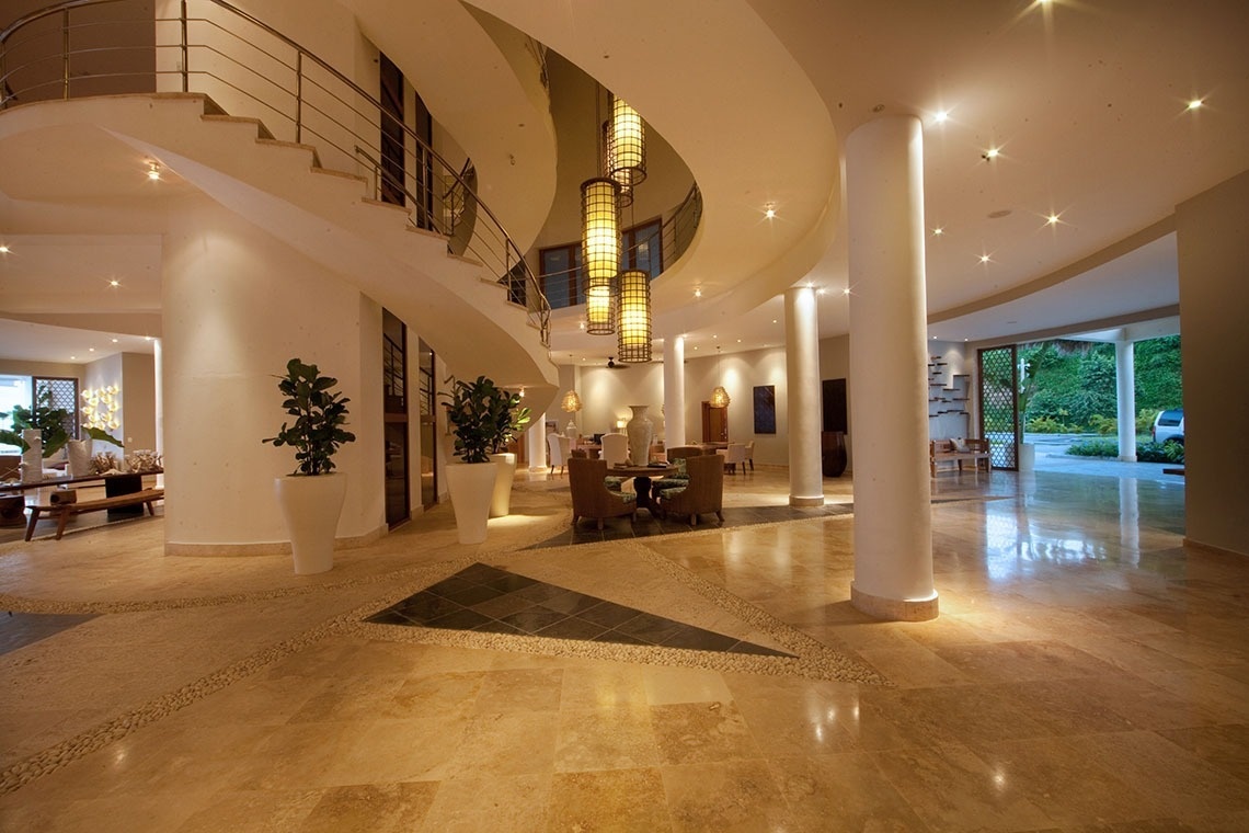 A spacious and elegant modern lobby showcases a dramatic spiral staircase, tall pillars, and warm, inviting light reflecting off its polished stone floor.