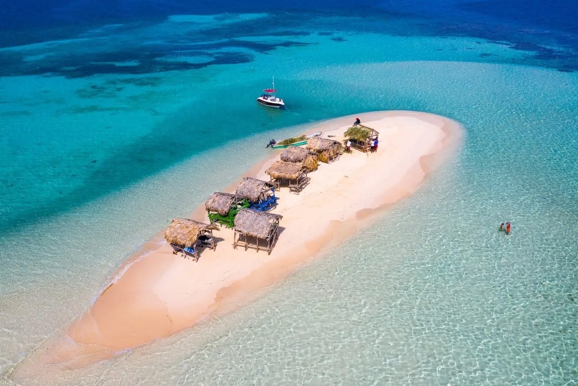An aerial view captures a small sandy island featuring several thatched huts, surrounded by vibrant turquoise and clear blue ocean water, with a boat anchored nearby and people wading in the shallows.