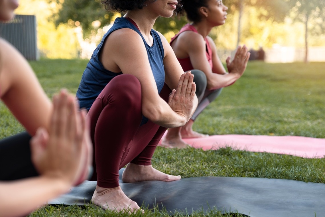 Three women are practicing yoga outdoors in a squatting position with their hands in a prayer pose.