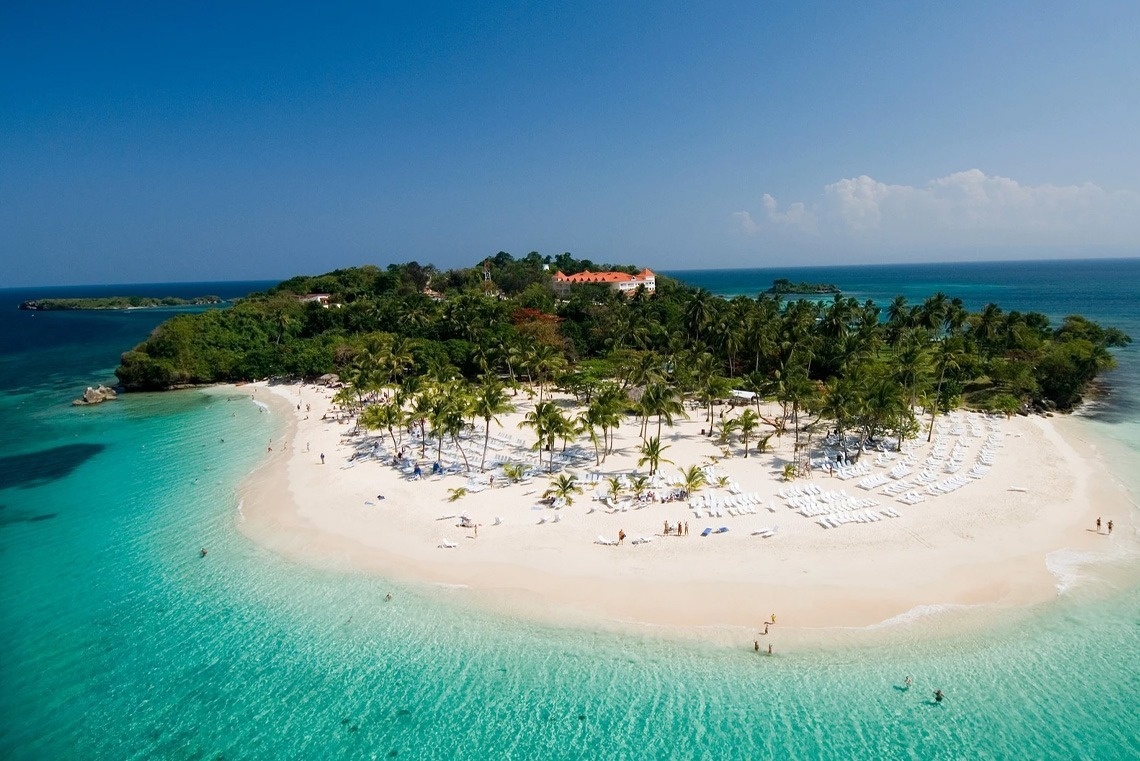 An aerial view showcases a picturesque tropical island with a white sandy beach, crystal-clear turquoise waters, and a building peeking through lush palm trees.