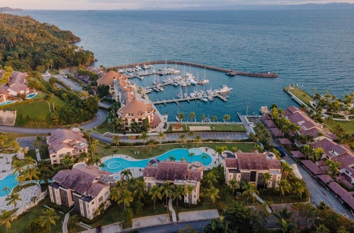 An aerial view captures a vibrant coastal resort featuring numerous buildings, multiple swimming pools, lush green landscapes, and a busy marina filled with yachts, all situated beside a calm bay with distant hills.