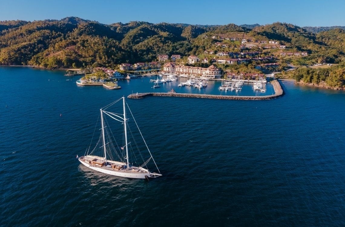 An aerial view captures a vibrant coastal resort featuring numerous buildings, multiple swimming pools, lush green landscapes, and a busy marina filled with yachts, all situated beside a calm bay with distant hills.