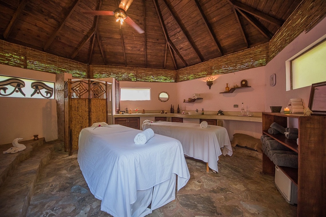 A rustic and serene spa room features two massage tables, a high wooden ceiling, stone floor, and decorative accents like bamboo screens and Eye of Horus window designs, creating a tranquil atmosphere.