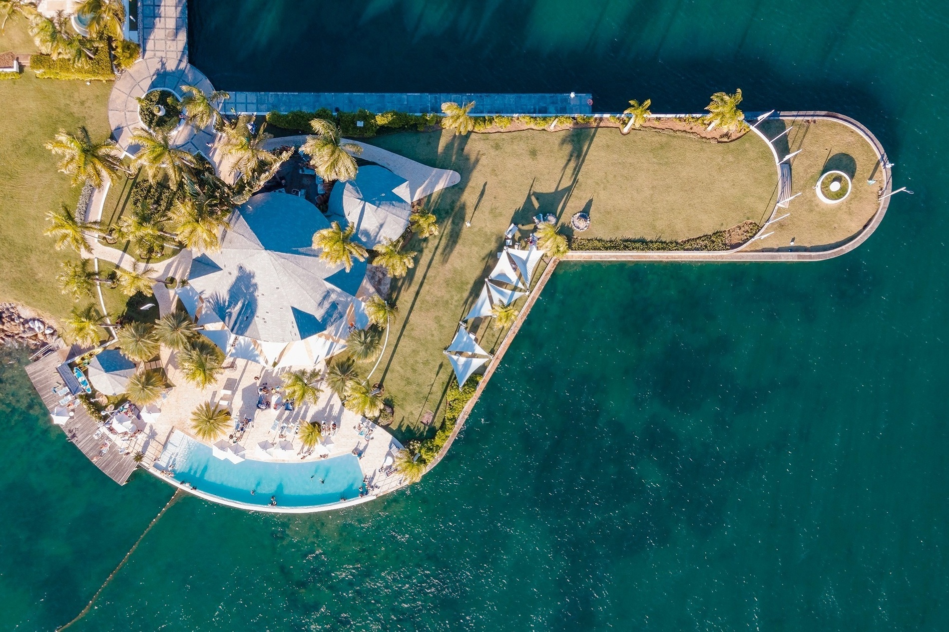 An aerial view captures a luxurious resort on a peninsula, featuring lush green lawns, numerous palm trees, a curving swimming pool, and paved areas, all surrounded by clear blue-green water.