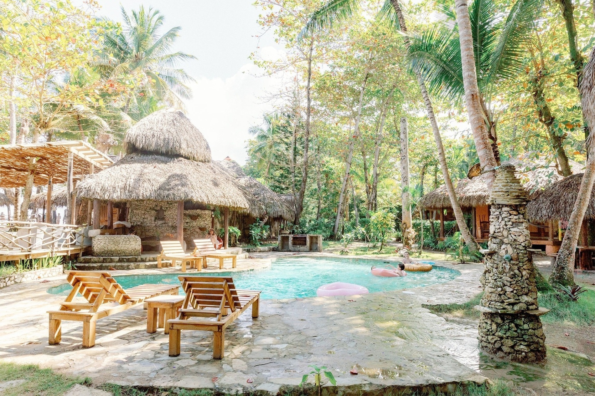 A vibrant tropical scene captures a swimming pool with people relaxing on inflatables, surrounded by rustic thatched-roof bungalows and abundant palm trees.