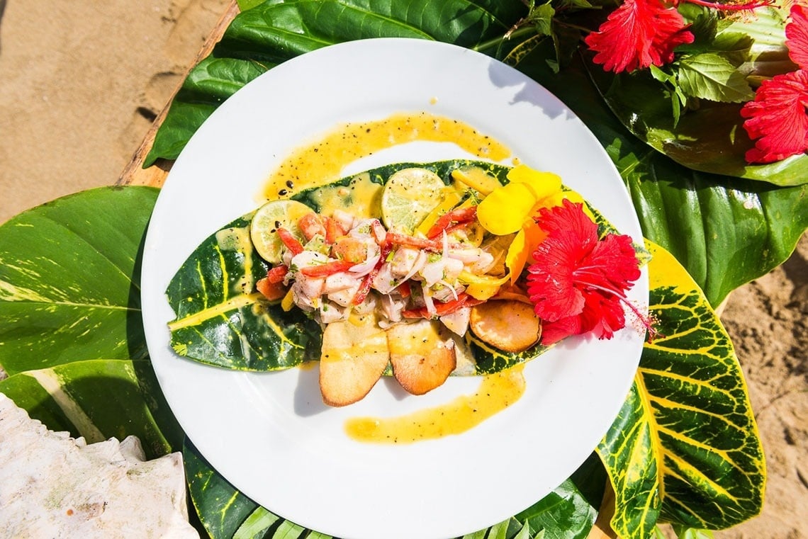 A vibrant tropical dish, possibly ceviche with shrimp and plantains, is artfully arranged on a white plate over large green and yellow leaves, garnished with red and yellow hibiscus flowers, and set against a sandy background.