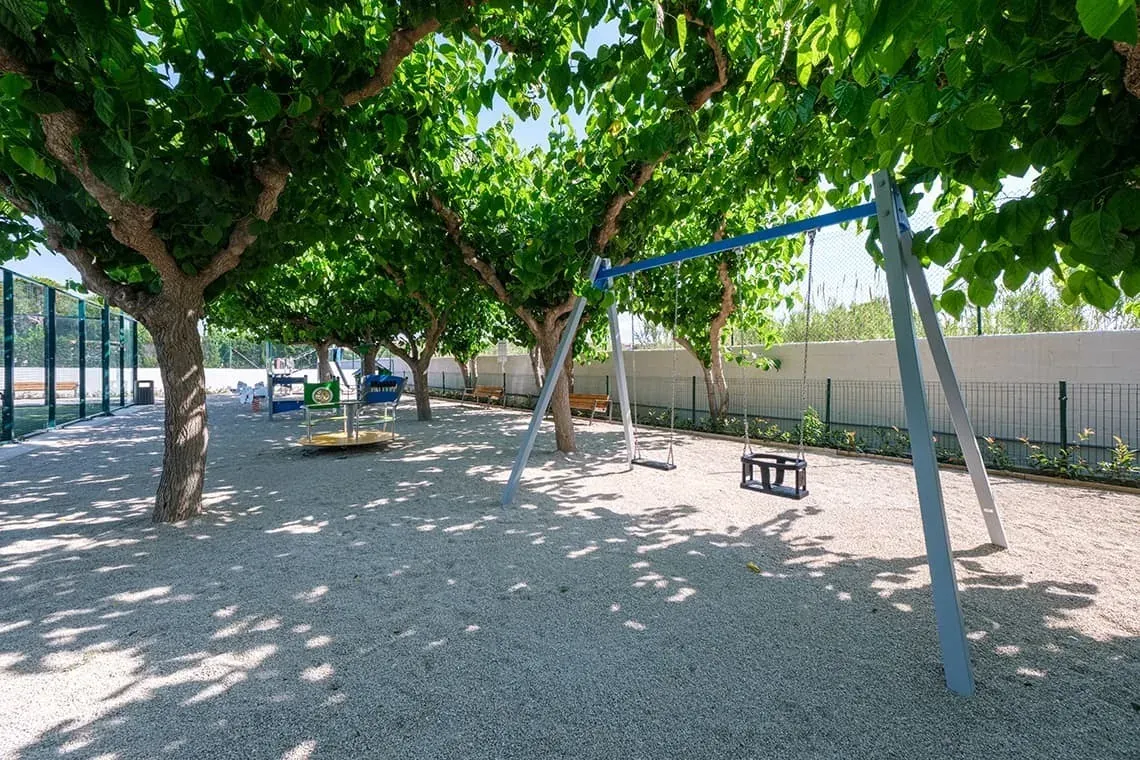 Parque infantil ESTIVAL: Corporativa, columpios y tiovivo bajo árboles frondosos.
