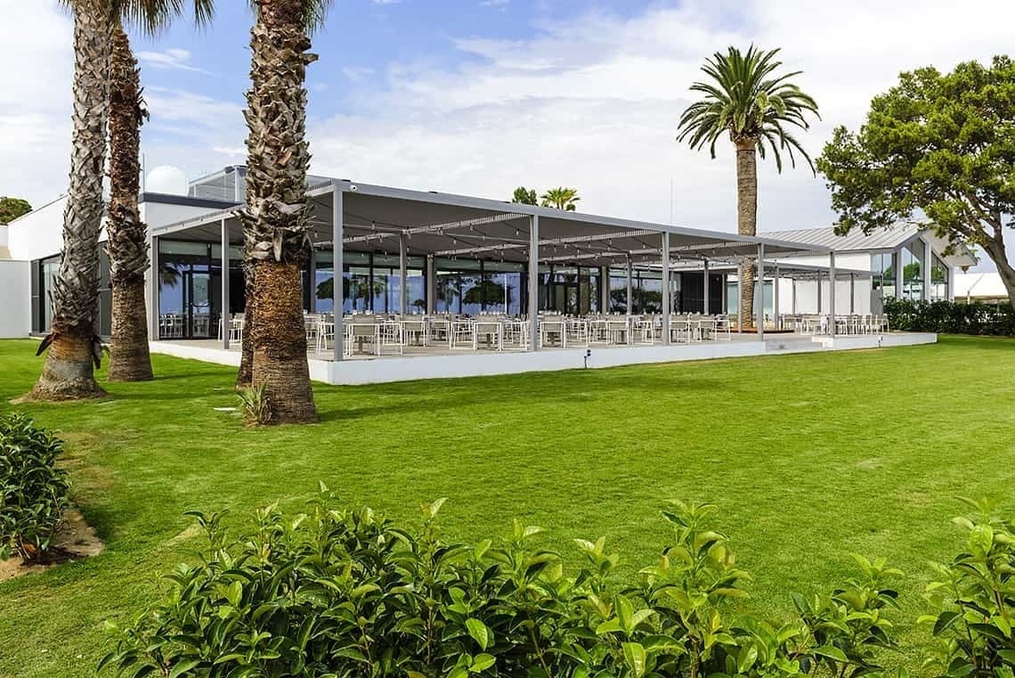 Estival Eldorado Resort: Amplio patio con mesas y sillas bajo un toldo, palmeras y césped verde. Ideal para eventos. Estival Group | Web Oficial.