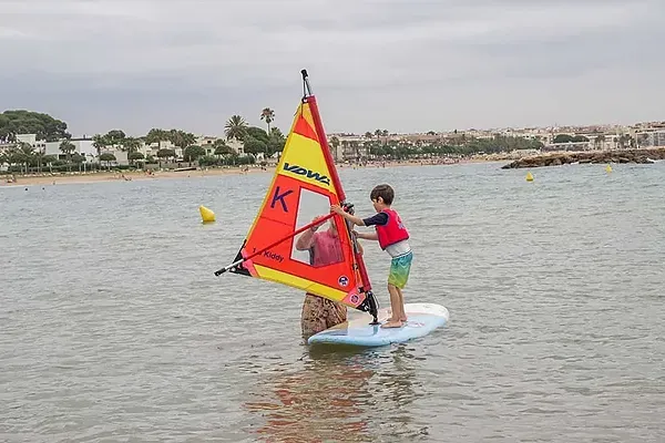 ESTIVAL: Corporative. Enfant apprenant la planche à voile en eaux calmes, plage en arrière-plan.