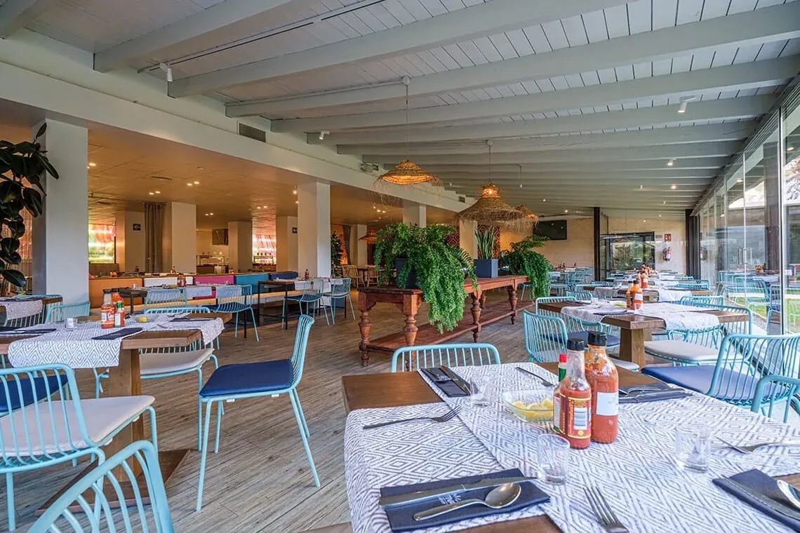 Salle à manger ESTIVAL: Corporativa, tables dressées, chaises bleues, ambiance tropicale.