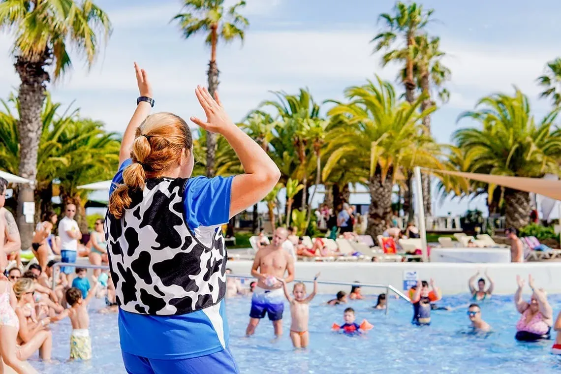 ESTIVAL: Animateur leitet Kinderaktivität im Pool, mit Palmen und entspannenden Gästen.