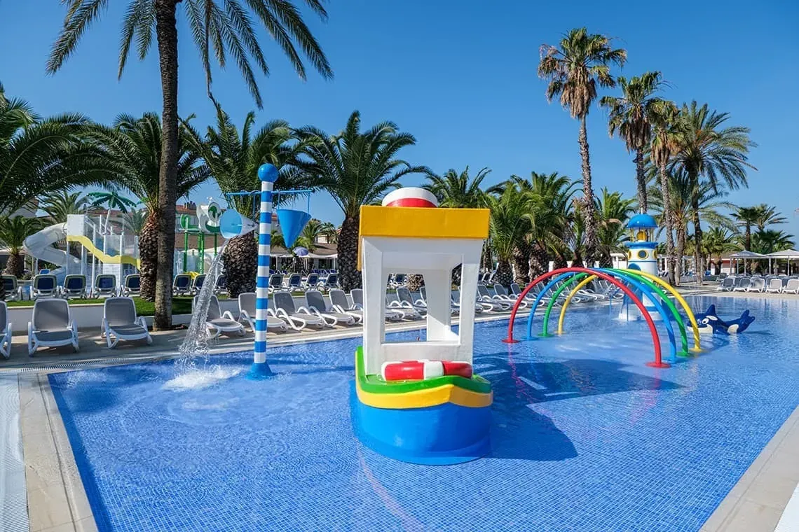 ESTIVAL: Piscina infantil con juegos acuáticos y palmeras para vacaciones familiares.