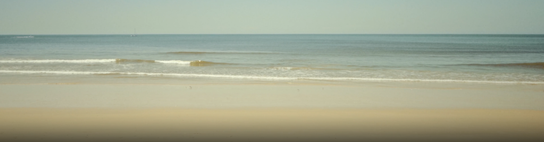 Uma paisagem serena de praia e oceano apresenta areia dourada, ondas suaves a quebrar na costa, um céu azul claro e algumas embarcações distantes na linha do horizonte.