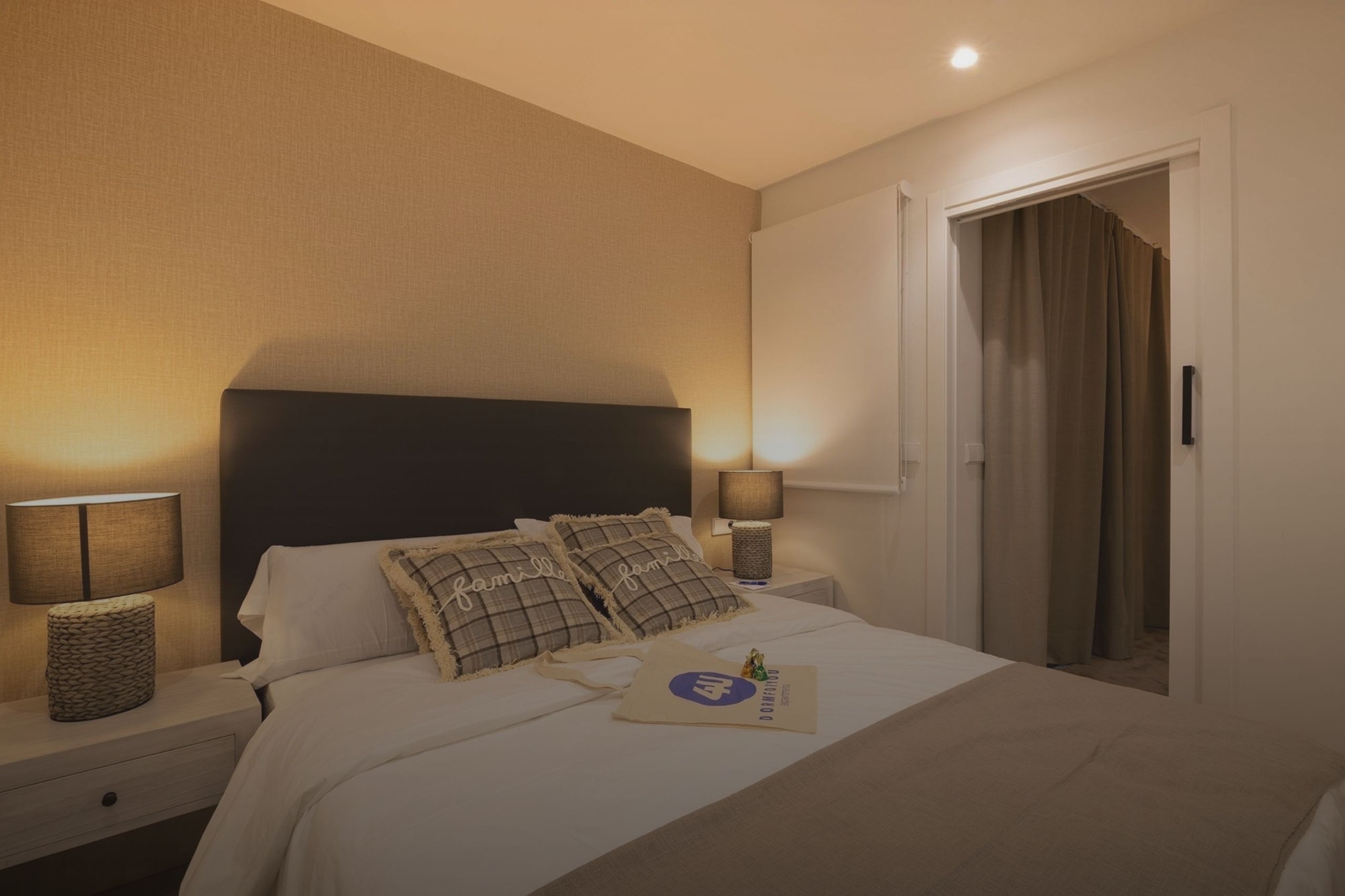 Un acogedor dormitorio presenta una cama hecha con almohadas decorativas, mesitas de noche con lámparas encendidas y una entrada con cortinas al fondo.