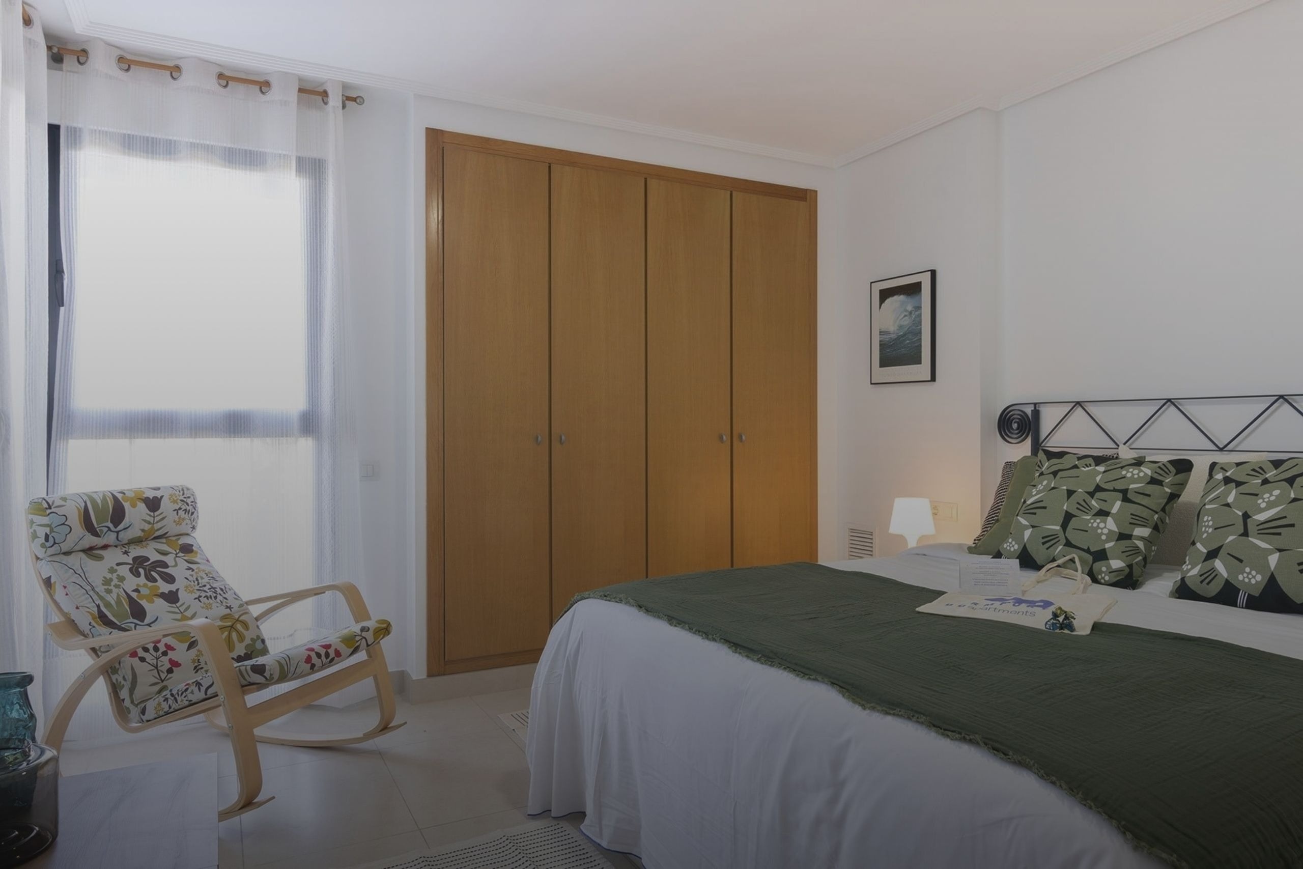 Un dormitorio bien iluminado presenta una cama con cabecero de metal y ropa de cama verde, un armario de madera empotrado, una mecedora con tapicería floral y una ventana con cortinas transparentes.