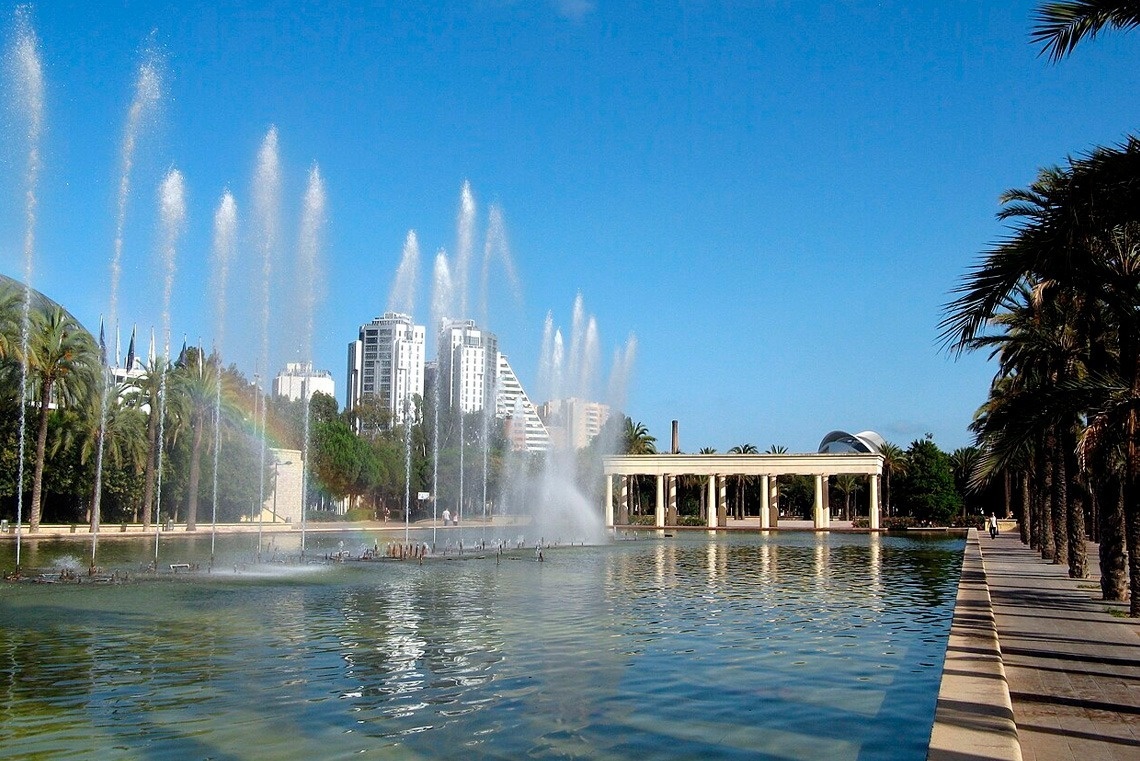 a fountain in a park with palm trees and buildings in the background