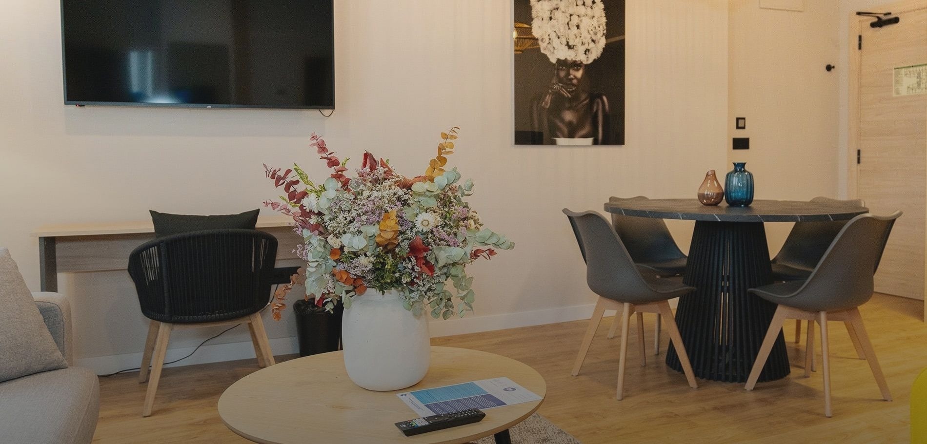 Una sala de estar y comedor moderna y luminosa cuenta con un gran arreglo floral vibrante en una mesa de centro de madera clara, un televisor de pantalla plana, y una mesa de comedor redonda oscura con sillas grises.