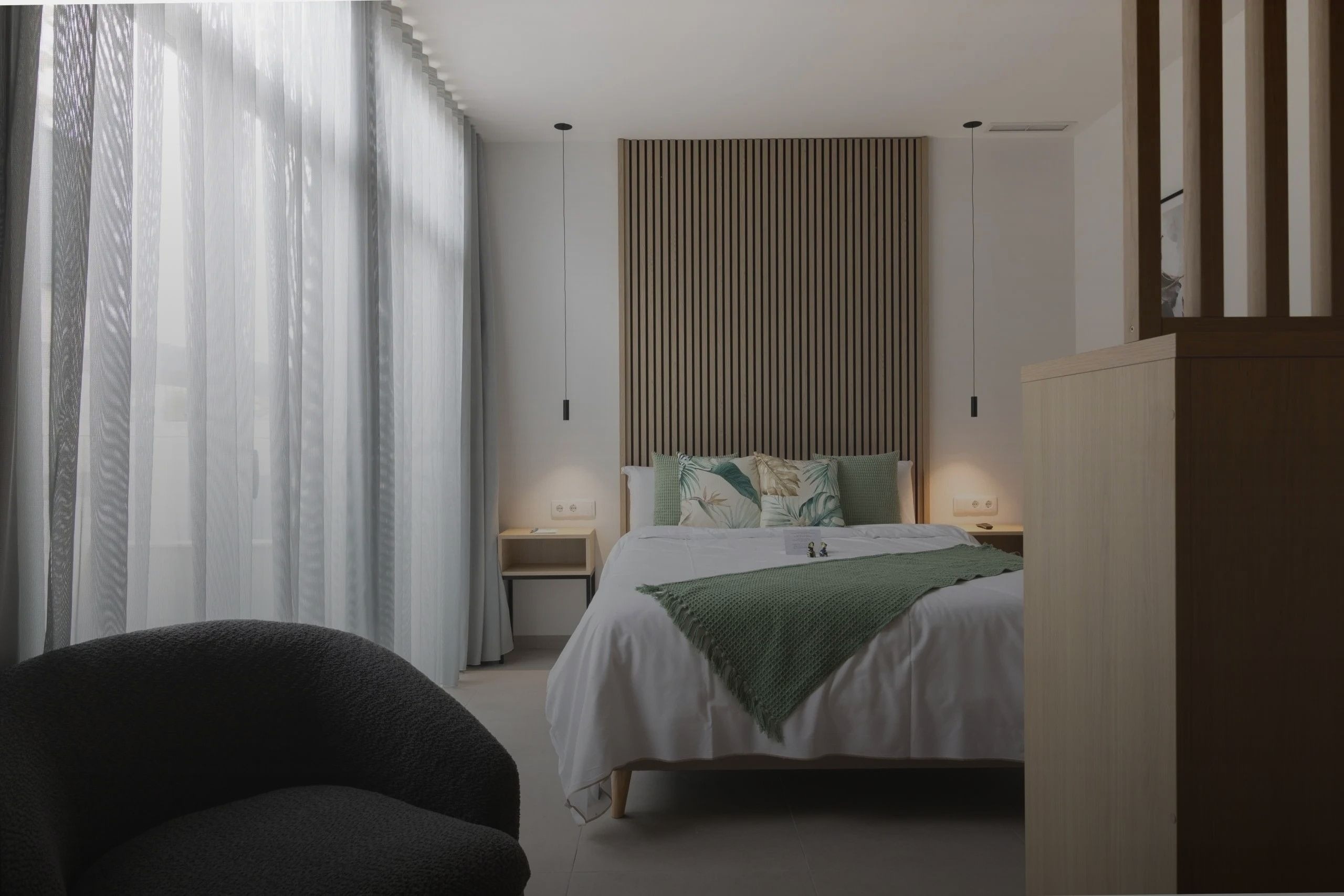 Una moderna habitación de hotel presenta una cama con ropa de cama blanca y detalles en verde, un cabecero de madera con lamas verticales y cortinas translúcidas que cubren una ventana.
