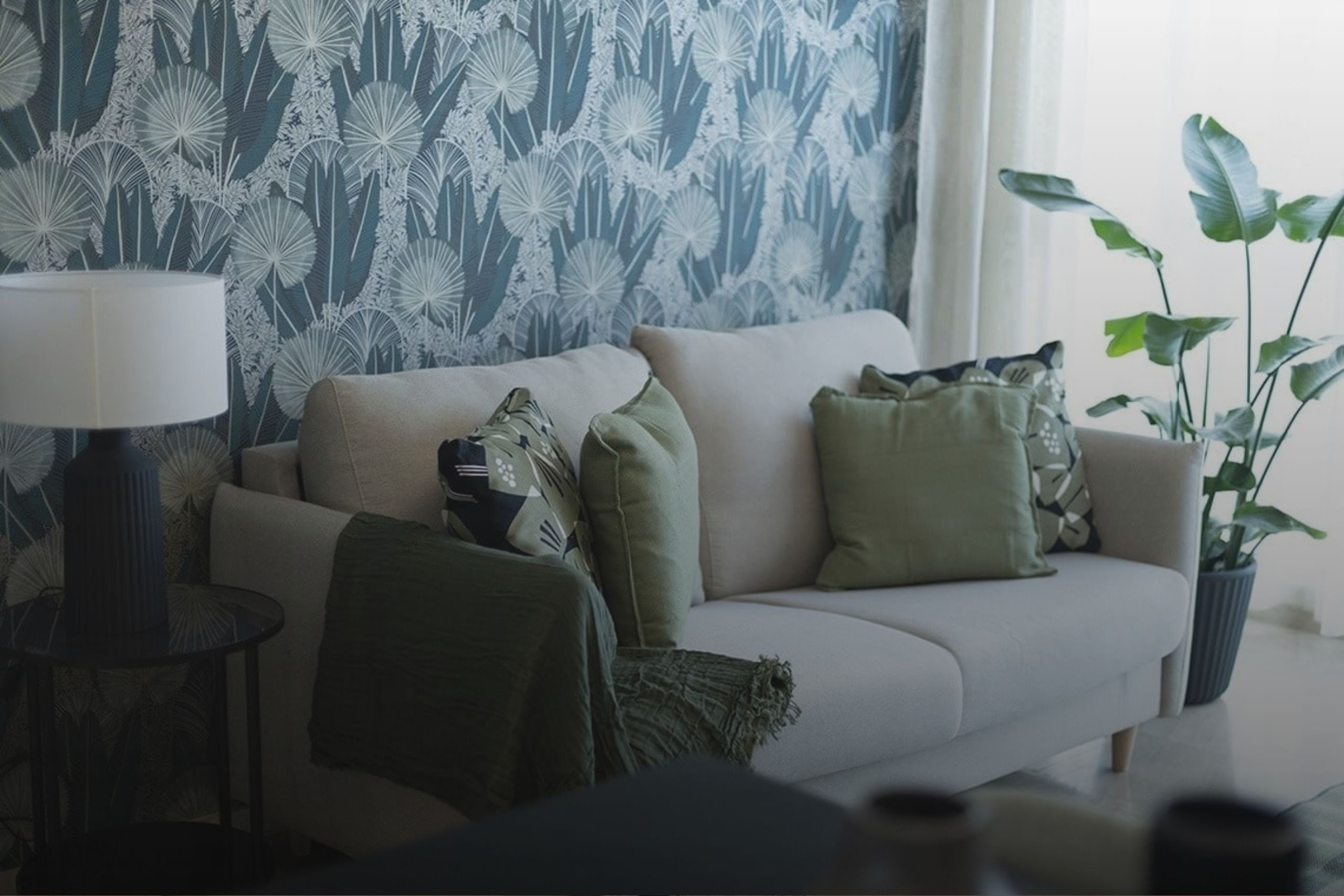 Un salón moderno exhibe un sofá beige con cojines y una manta verdes, enmarcado por una pared con papel pintado de hojas de palma azul verdoso, una lámpara de mesa y una gran planta.