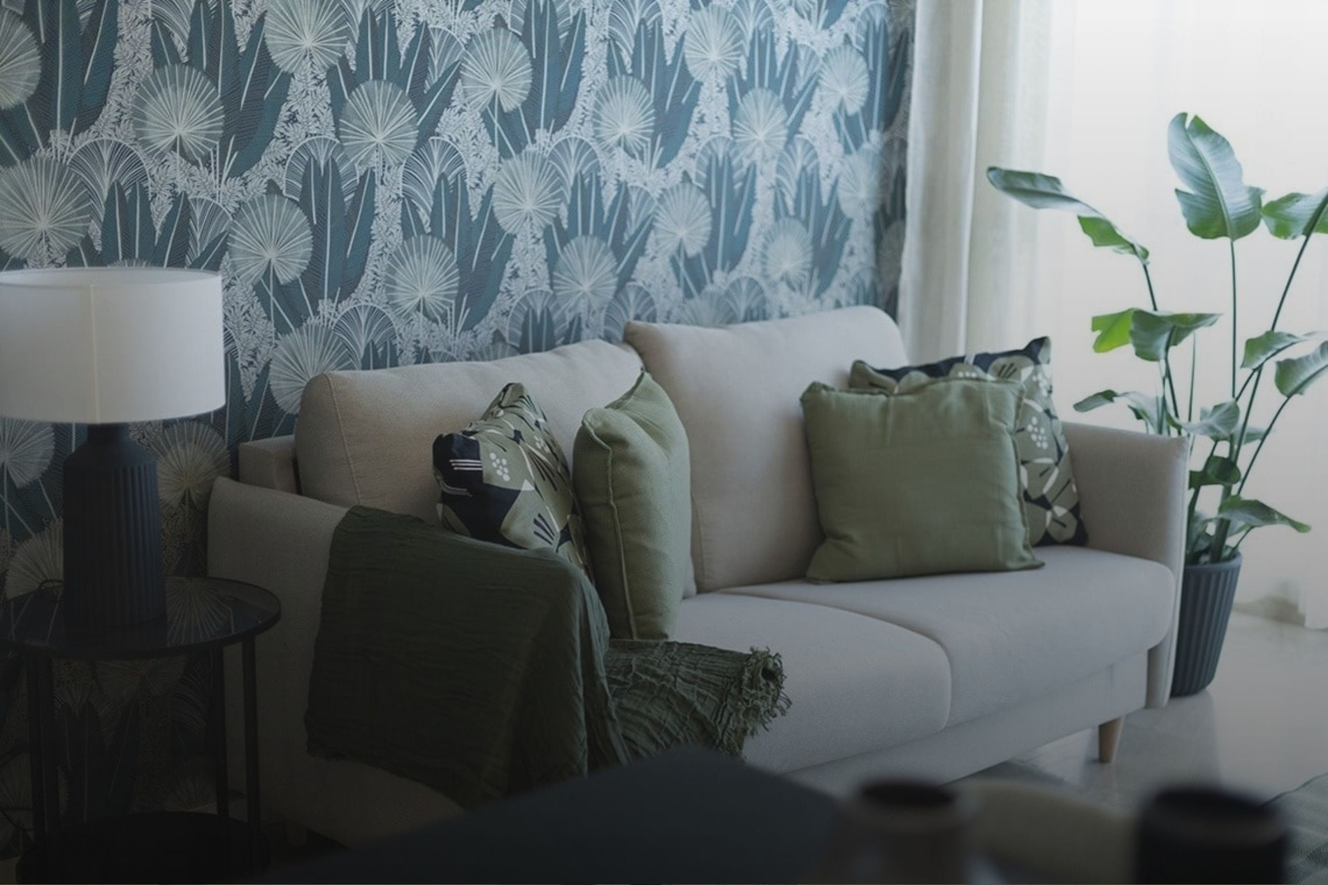 Un salón moderno exhibe un sofá beige con cojines y una manta verdes, enmarcado por una pared con papel pintado de hojas de palma azul verdoso, una lámpara de mesa y una gran planta.