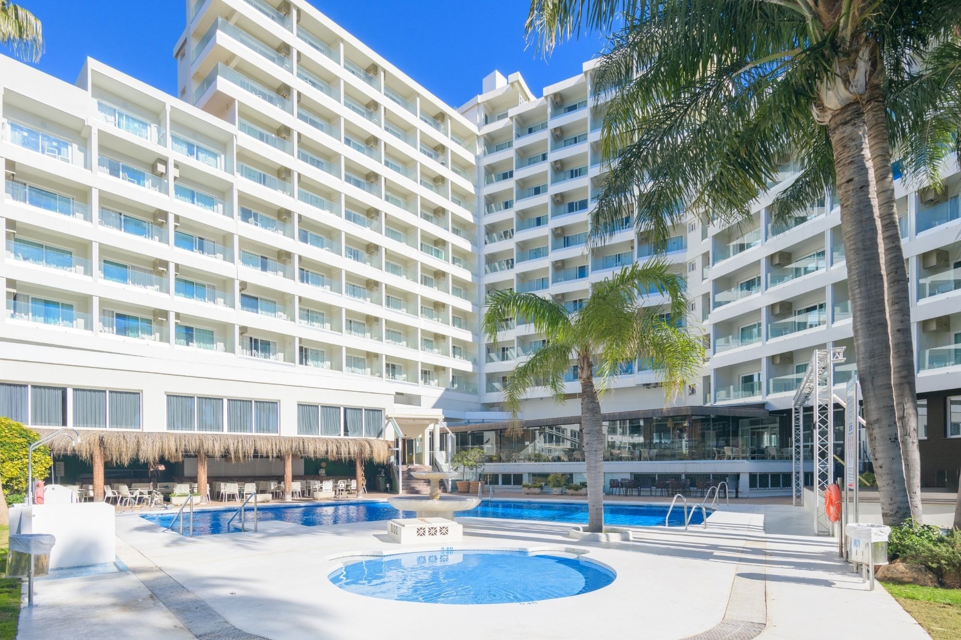 A large, white hotel building with numerous balconies overlooks a bright blue swimming pool area, complete with a small, circular pool, and is surrounded by lush palm trees under a clear sky.