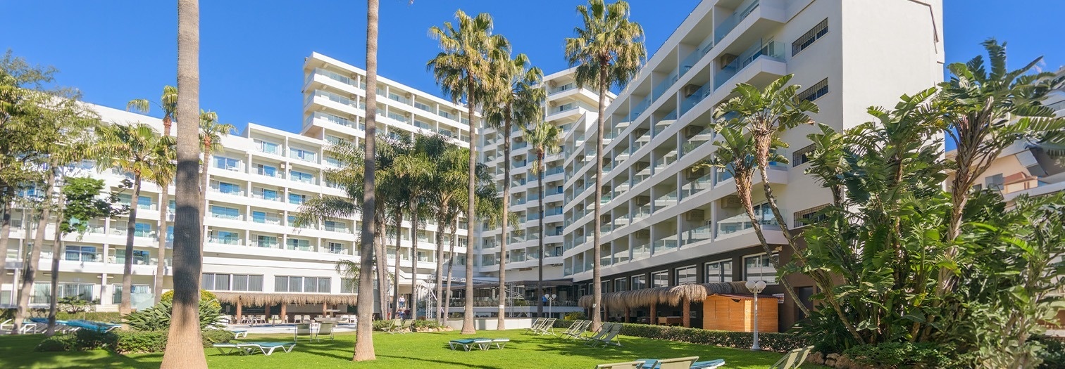 Un hotel blanco con balcones y palmeras se erige sobre un césped verde con tumbonas, bajo un cielo azul.