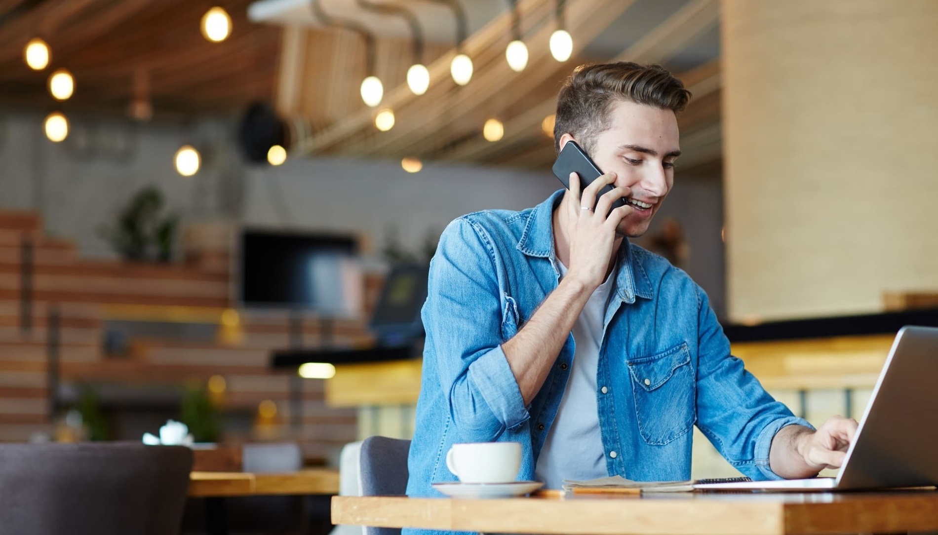 um homem está sentado em uma mesa usando um laptop e falando ao celular