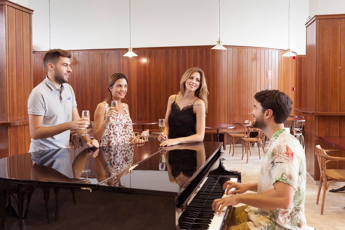 um grupo de pessoas está sentados ao redor de um piano enquanto um homem toca