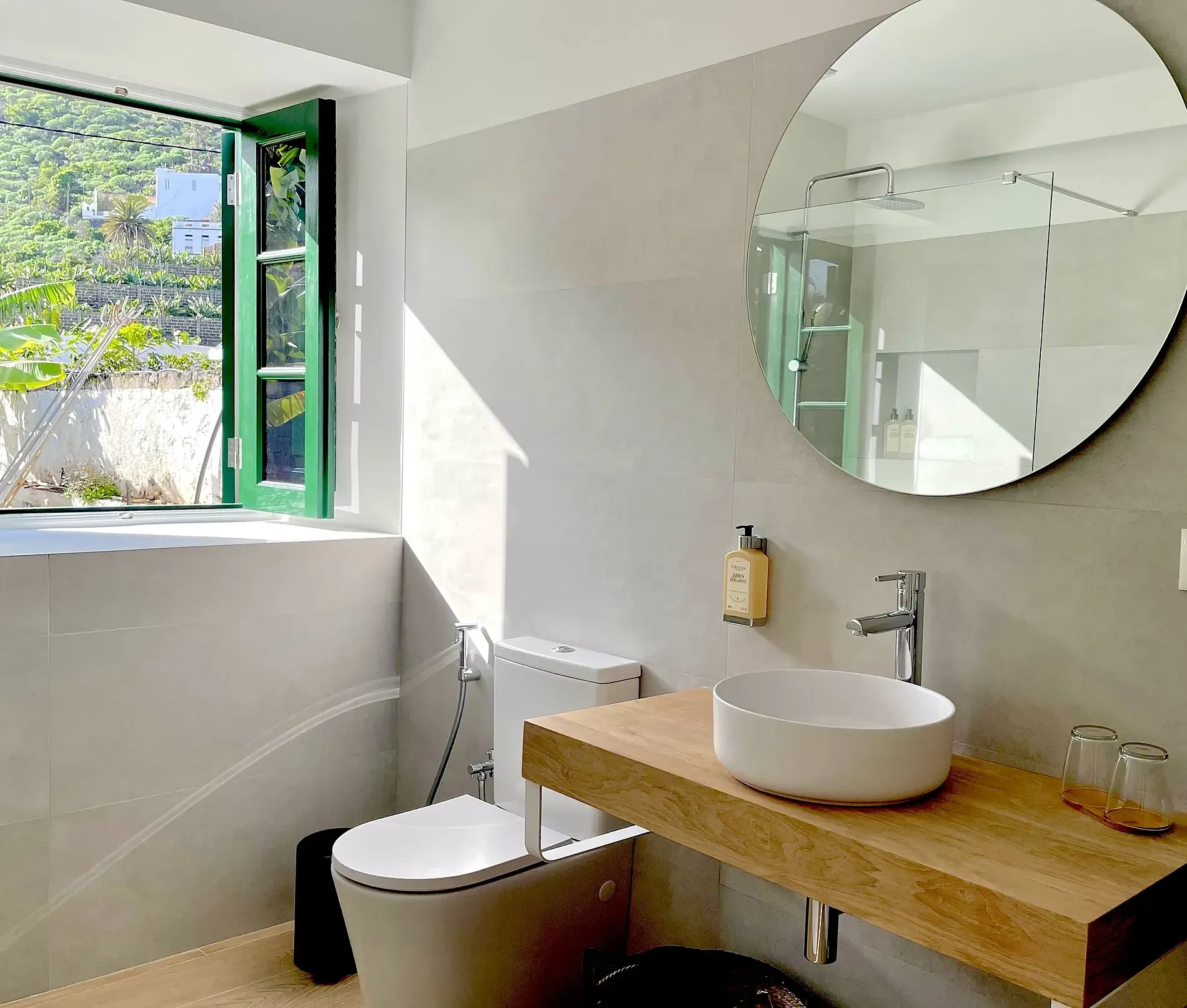 Baño moderno con ventana verde, ducha con espejo circular y lavabo sobre encimera de madera. El patio