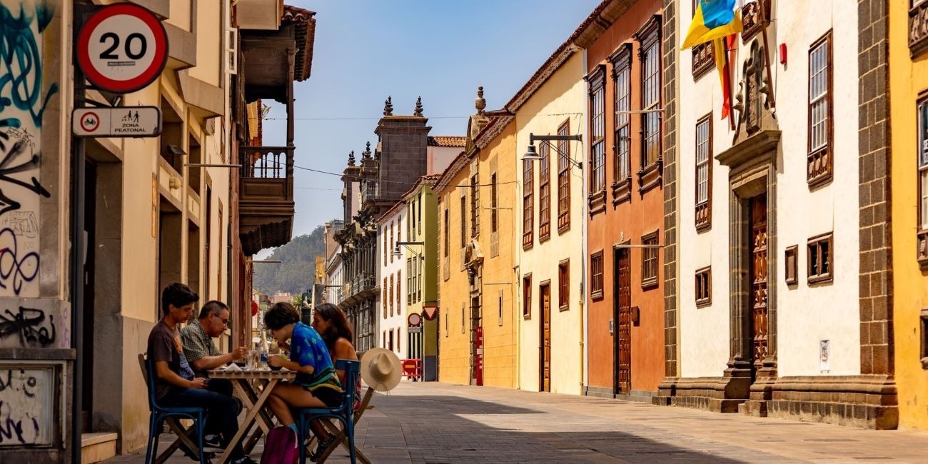 Una calle europea vibrante y soleada presenta edificios de fachadas coloridas mientras un grupo de personas disfruta de una comida al aire libre en una terraza.