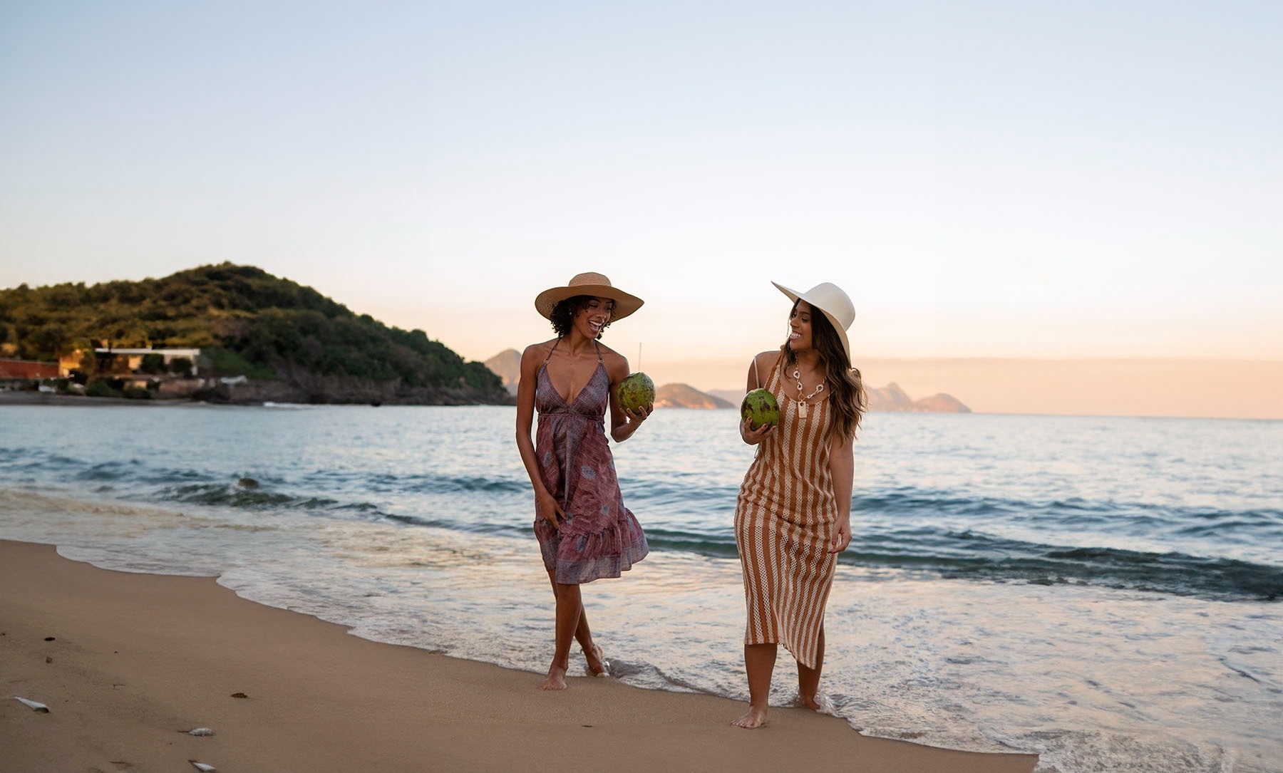 two women walking on the beach holding coconuts