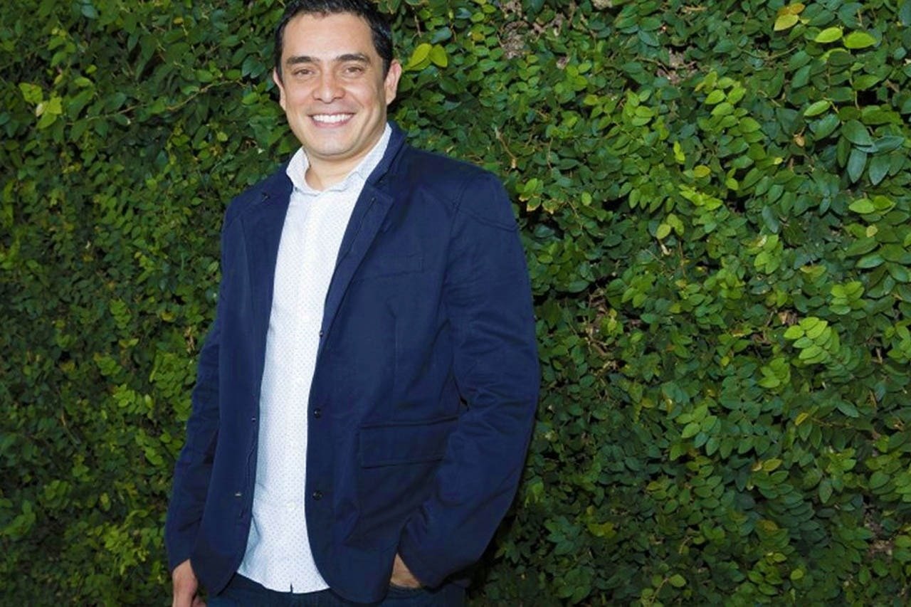 um homem de terno azul está parado em frente a uma sebe verde