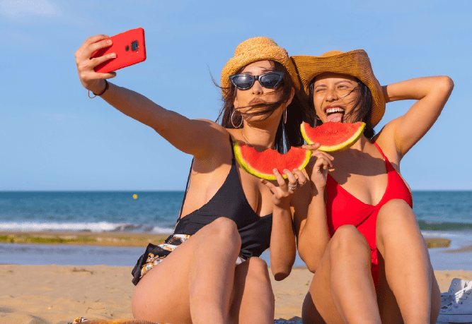 two women are eating watermelon on the beach and taking a selfie